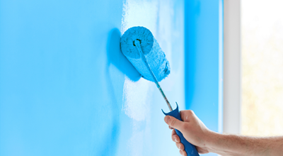 Hand using a paint roller to apply bright blue paint to a wall near a window.