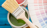 Paintbrush on an open can of green paint, with color swatches and another paint can in the background.