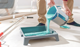 Person pouring light blue paint into a grey paint tray on the floor.