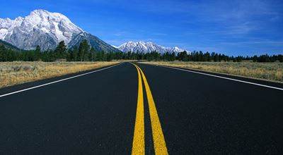 Freshly paved road set against pine trees and snow-topped mountains