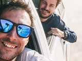 two young men leaning out from automobile windows, one with sunglasses that reflect his arm holding the camera taking the picture.