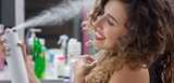 Smiling woman with curly hair sprays product from a white canister in a room with toiletries behind her.