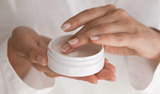 Hands holding a white cosmetic jar, with a finger dipping into the cream inside.