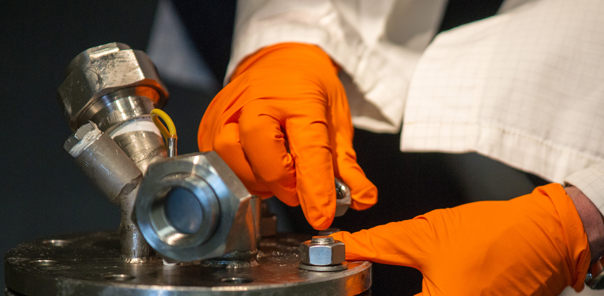 Person wearing orange gloves working with a metal bolt on industrial equipment.