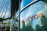 Glass building facade with the Nouryon logo, reflecting trees and a blue sky with clouds.