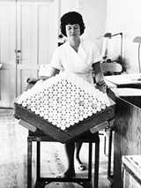 Woman in a white uniform stands behind a large tray holding neatly stacked candles at factory Liljeholmens Stearinfabrik in Stockholm.