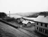Black and white photo of riverside industrial buildings in Stockholm with a chimney, fences, and houses near the water.
