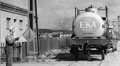 A worker holding a flag guides an EKA Bohus chemical tanker train on tracks in an industrial area.