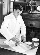 A man with a white coat spreads casein glue on wood with a tool in a factory.