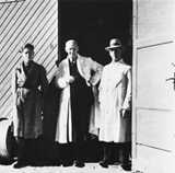 Three men in long coats stand outside a building.