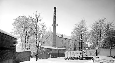 Factory building with a tall chimney labeled "CASCO" in a snowy landscape surrounded by trees and fences.