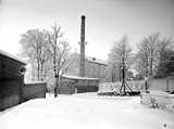 Factory building with a tall chimney labeled "CASCO" in a snowy landscape surrounded by trees and fences.