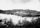 Large industrial factory complex with tall smokestacks by the water, surrounded by trees and hills.