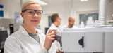 Woman in a white lab coat and safety glasses operates equipment in a laboratory, with colleagues in background.