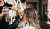 Hair stylist spraying hairspray on a woman's styled hair in a salon setting.