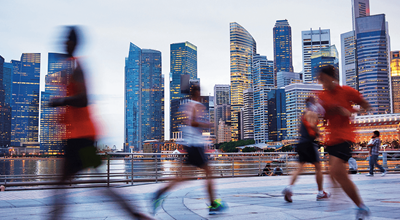 Faded people jogging along a waterfront promenade in the foreground with a city skyline and tall buildings in the background.